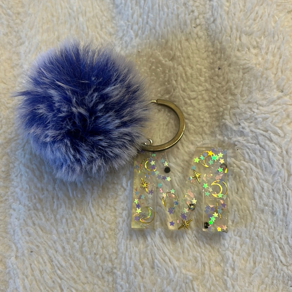 A keychain that has the initial "M" with a fuzzy ball attached to it